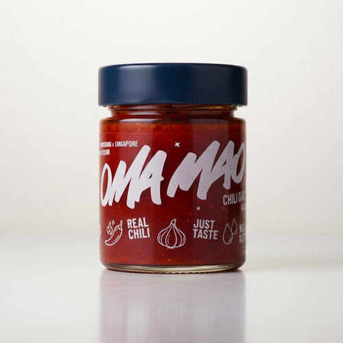 A jar of Oma Mao Chili Sauce on a light-colored tabletop with a blurred indoor background. The product features a dark blue lid and a red label with white text and illustrations of a chili pepper and garlic bulb.